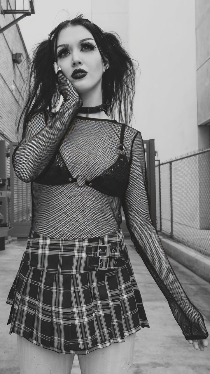 Model wearing a black long-sleeve fishnet top over a black bra with a plaid skirt.