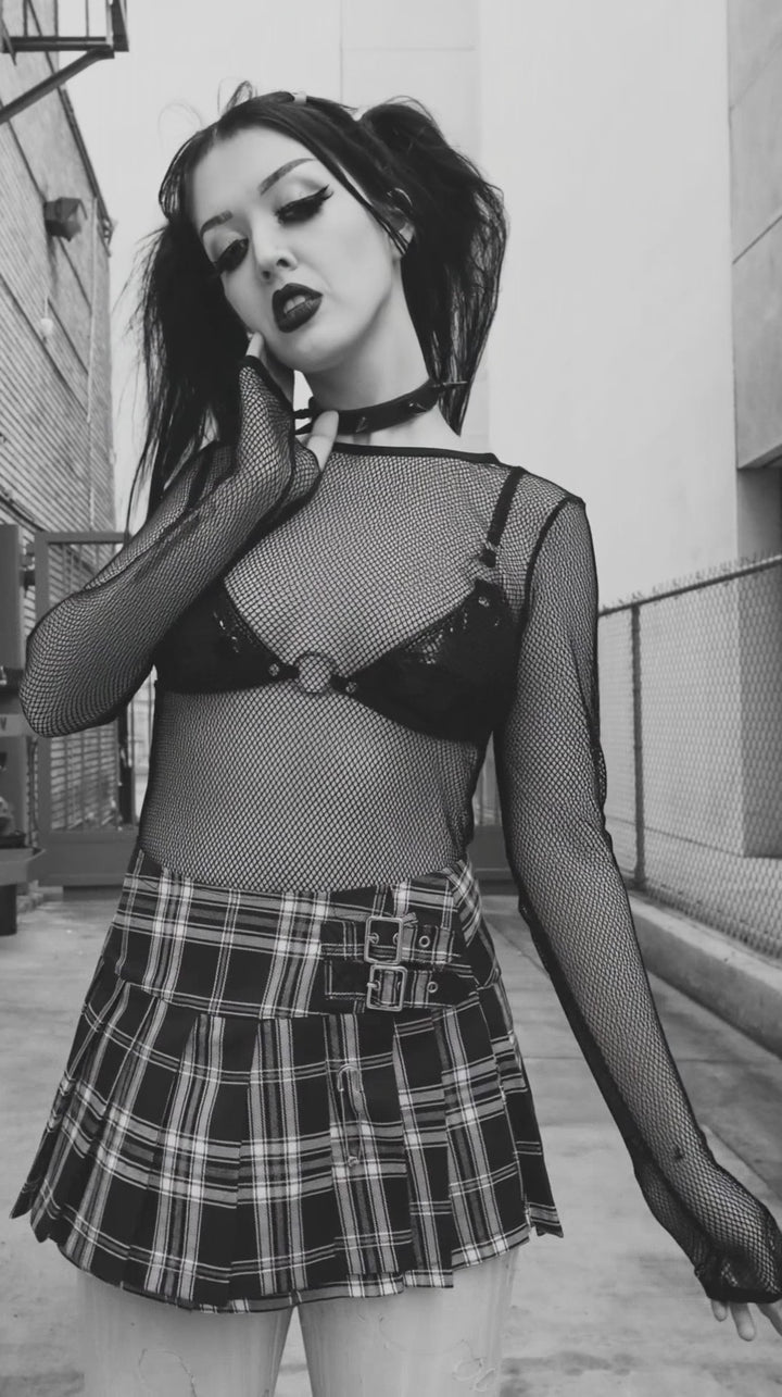 Model wearing a black long-sleeve fishnet top over a black bra with a plaid skirt.